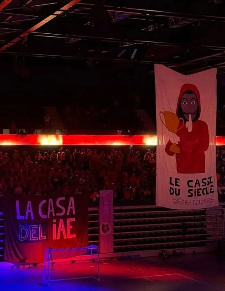 TIFO suspendu IAE 2025 en salle : masque La Casa de Papel tenant un trophée, suspendu à la verticale, bâche La Casa del IAE en contrebas